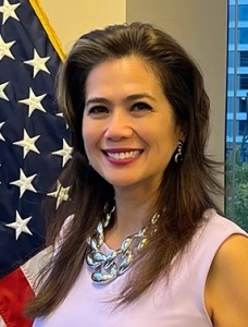 A smiling woman with long brown hair wearing a pink shirt and silver necklace poses in front of a U.S. flag.