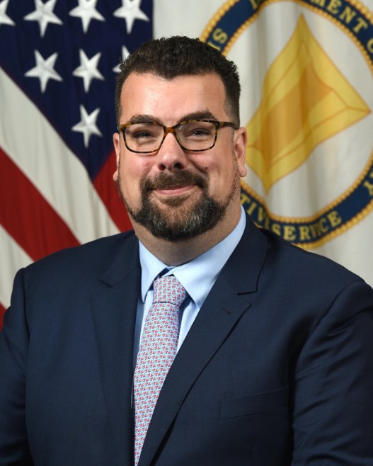 A man with short black hair and goatee and wearing glasses and a suit slightly smiles in front of a U.S. flag and another flag.