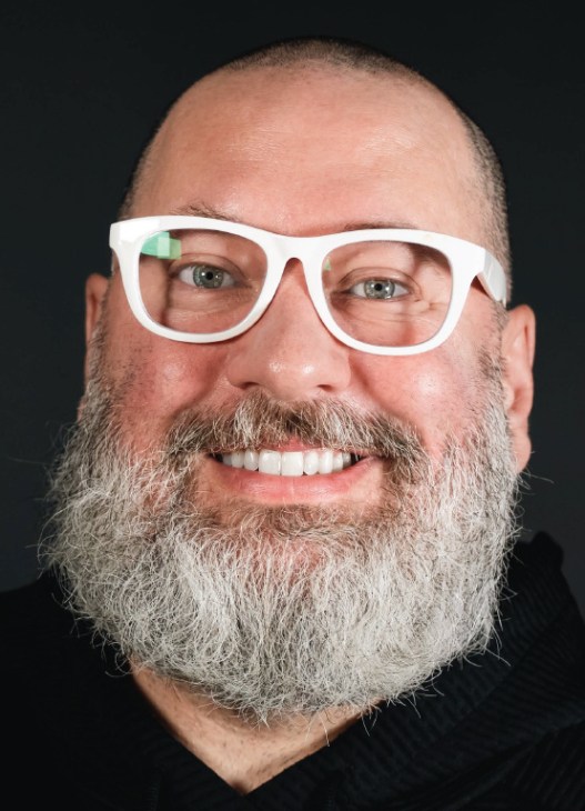 A smiling man with a large white beard wearing white-rimmed glasses and a black hoodie.