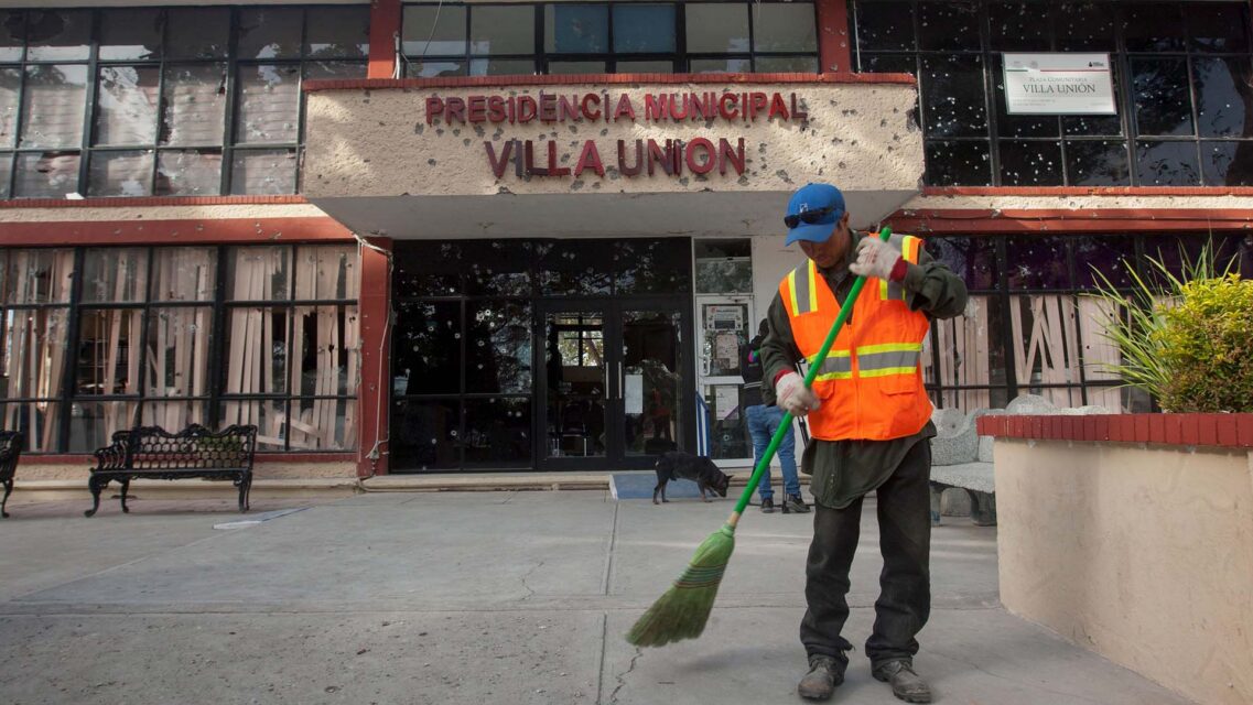 Photo of a worker sweeping the ground outside a building full of bullet holes with a sign that reads Presidencia Municipal Villa Union