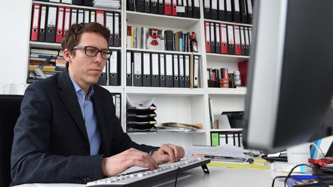 Journalist Bastian Obermayer sits at a computer keyboard at a desk in front of a bookshelf.