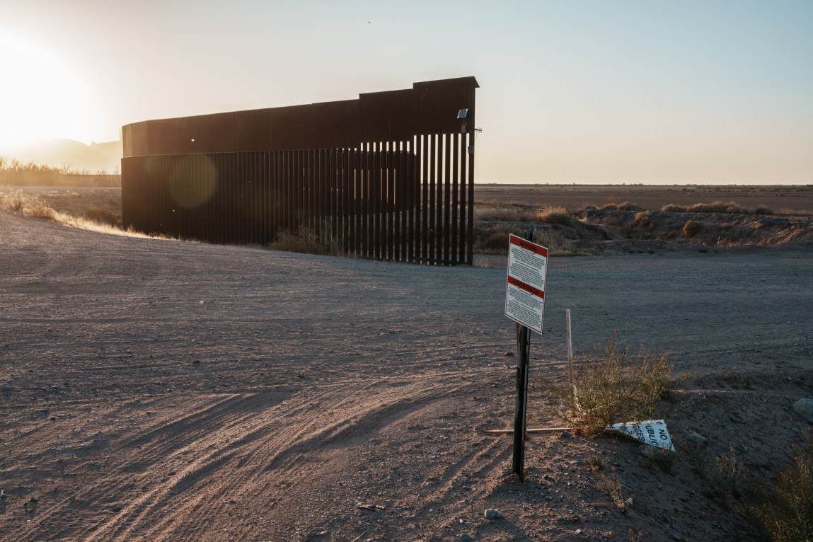 A sign on a post stating that the area is a military zone in English and Spanish, in front of a dirt road leading past an end point of a slotted steel bollard U.S. border wall with Mexico with a low, glowing sun in the distance.