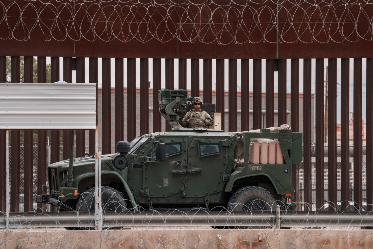 A U.S. Army vehicle in front of the slotted steel bollard border wall with Mexico, with razor wire hanging off it.