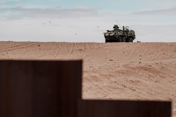 A U.S. Army vehicle sits parked on a sandy hill, with a small portion of the U.S. border wall blocking the scene.