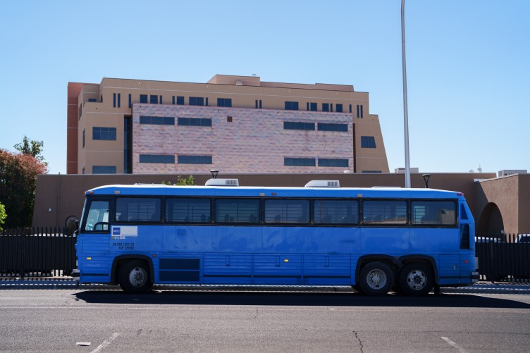 A large blue bus parked in a lot in front of a large tan building.