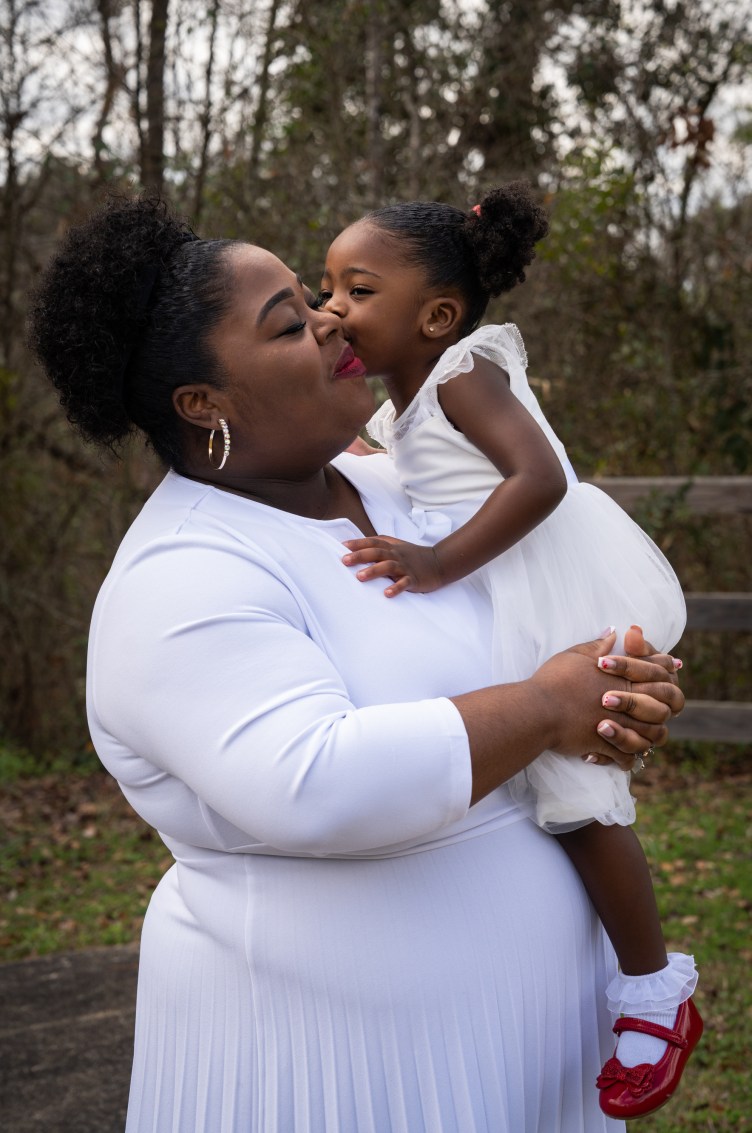 Outside, a Black woman in a white dress closes her eyes and smiles while holding a toddler wearing a white dress, white ruffled socks, shiny red shoes and pigtails.