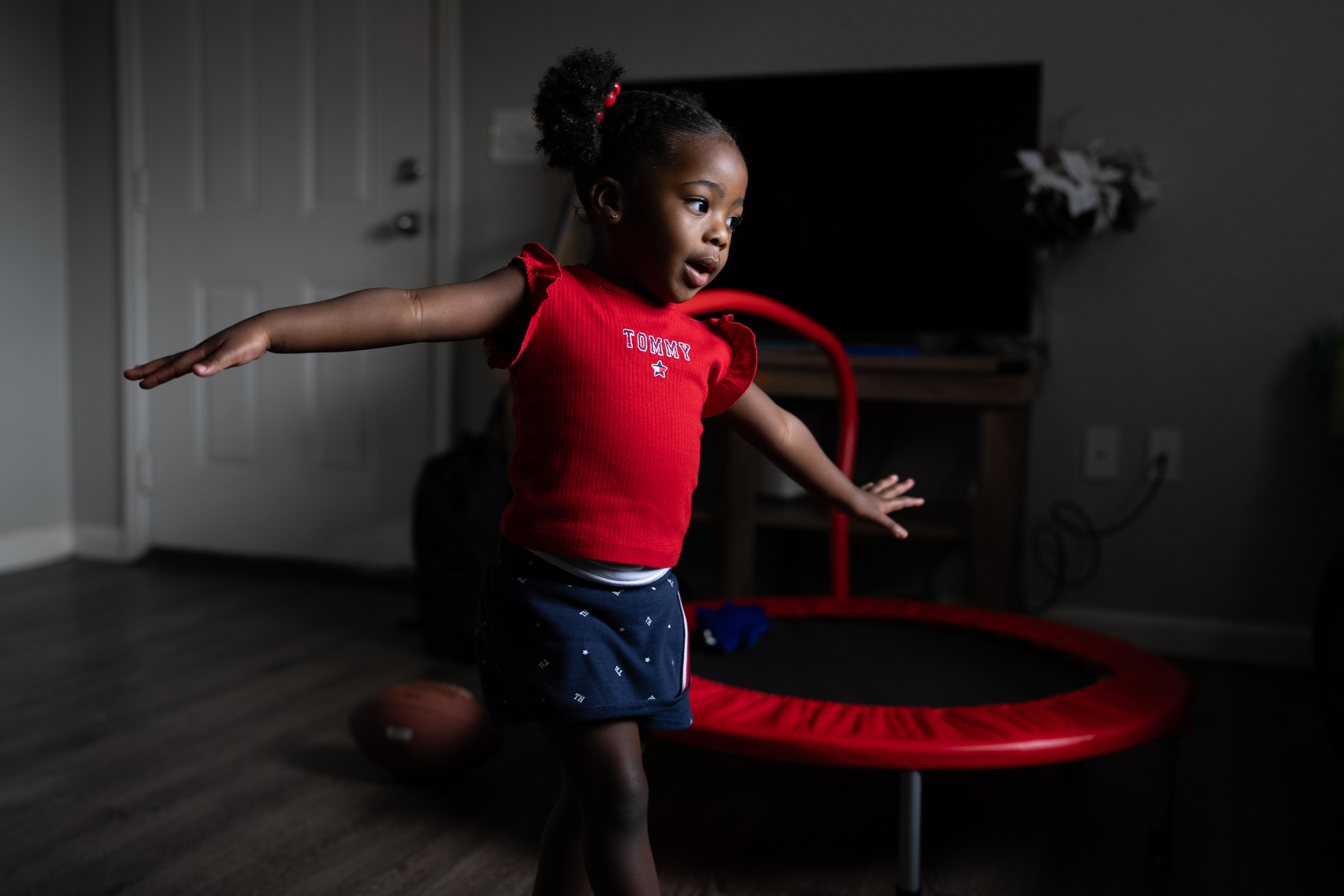 A child wearing a red shirt and blue shorts runs with her arms out past a red trampoline, a football and a television set inside of a dimly lit, tidy room.