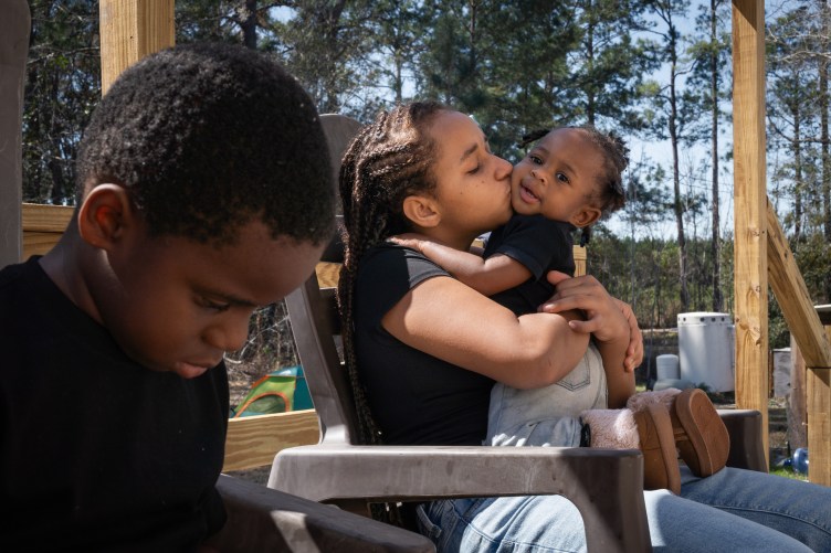 Three children sit outside in plastic chairs in an open wooden structure. In the background are trees and a camping tent. One of the children holds a baby in her arms and kisses her.