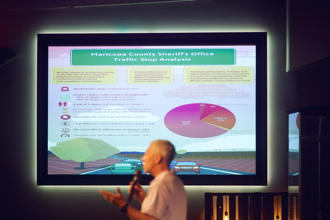 A man speaks into a microphone in a dark room. A projection screen behind him displays the headline “Maricopa County Sheriff’s Office Traffic Stop Analysis” above a pie chart and a number of bullet points, but the text is too small to read.