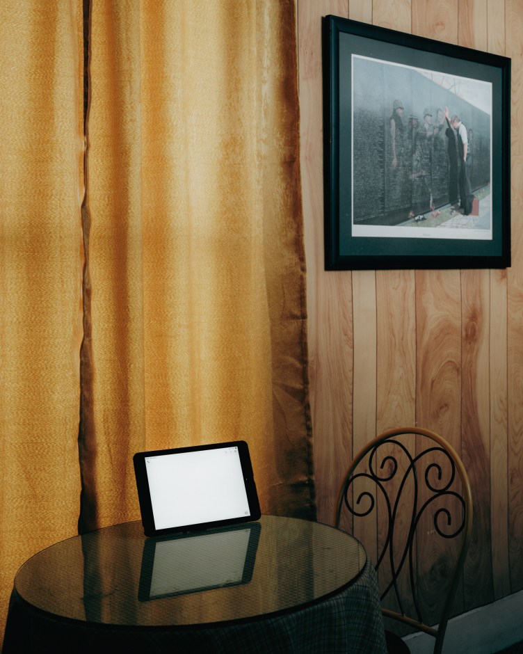 A glowing iPad screen sits on a small glass kitchen table near a window covered with a gold-colored curtain and wood-panneled walls.