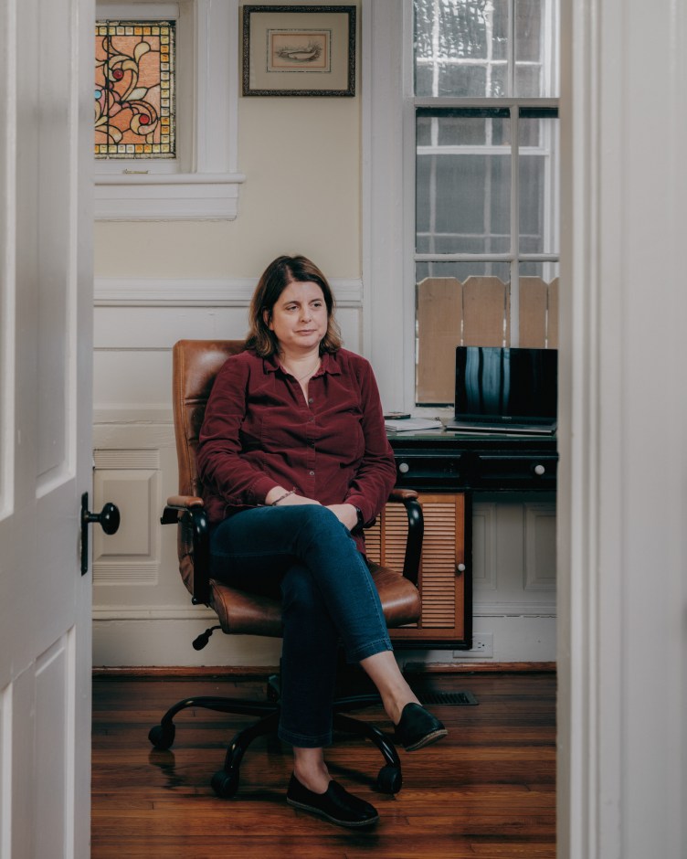 A woman wearing a maroon button-up shirt and blue pants sits in an office chair near a desk with a laptop and notepad.
