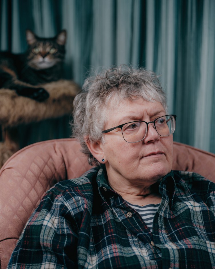 A woman with short graying hair, wearing glasses and a checkered shirt, sits on a pink sofa with a cat and blue curtain behind her.