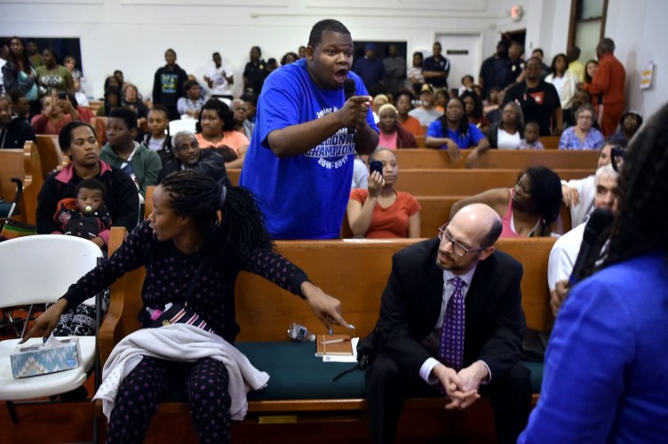 A man yelling into a microphone points a finger at other speakers, in a church where dozens of people are sitting in pews.