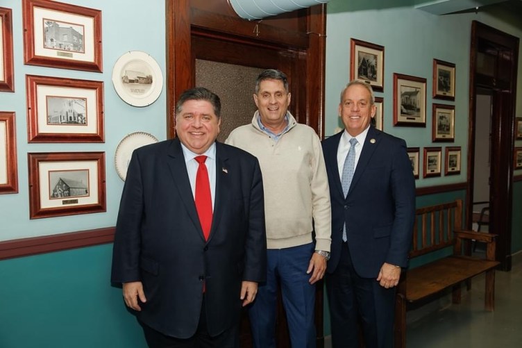 Three men in business attire look at the camera and smile, in a room with numerous framed black-and-white historical photos hung on the wall.