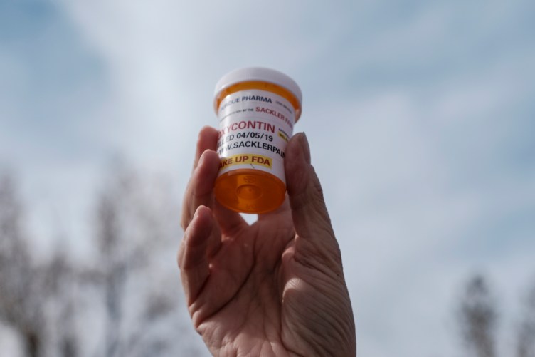 A hand holds a pill bottle in the air. The bottle says, “Purdue Pharma,” "OxyContin” and “Sackler.”
