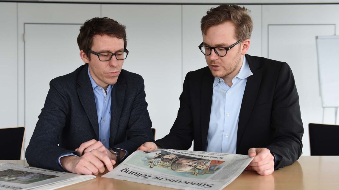 Bastian Obermayer and Frederik Obermaier sit at a desk with a Sueddeustche Zeitung newspaper spread out before them.