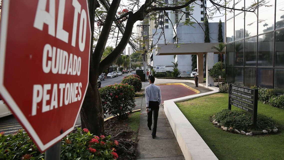 A man walks along a path outside a building between a red stop sign and a building nameplate that reads Mossack Fonseca
