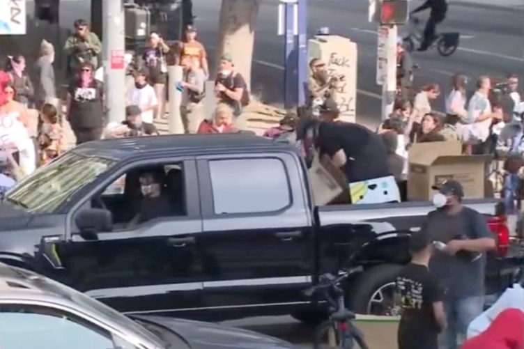 A man wearing a face mask sits in the driver’s seat of a truck with cardboard boxes in the bed. People are surrounding the truck and one person is crouched in the bed going through the boxes.