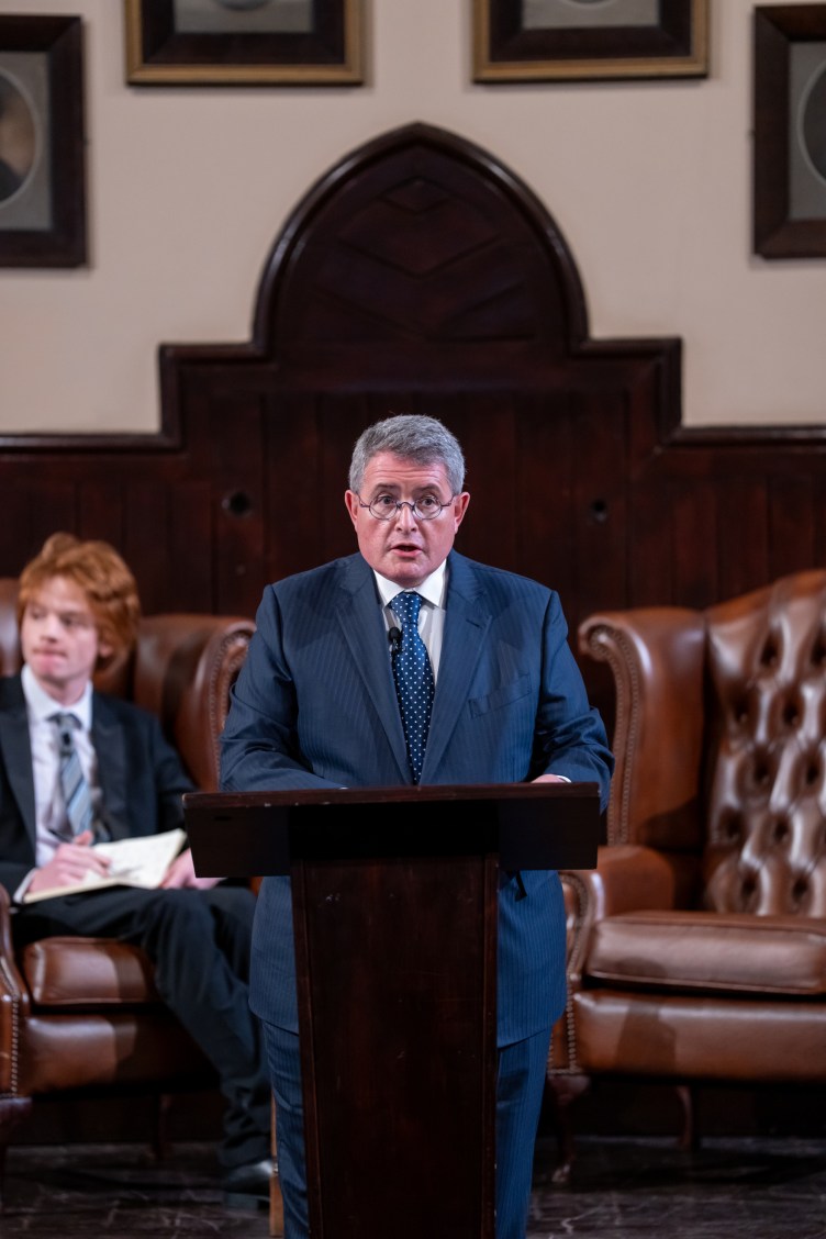 A man in a suit and tie stands behind a lectern with large leather armchairs and wood paneling behind him. He is speaking into a microphone.