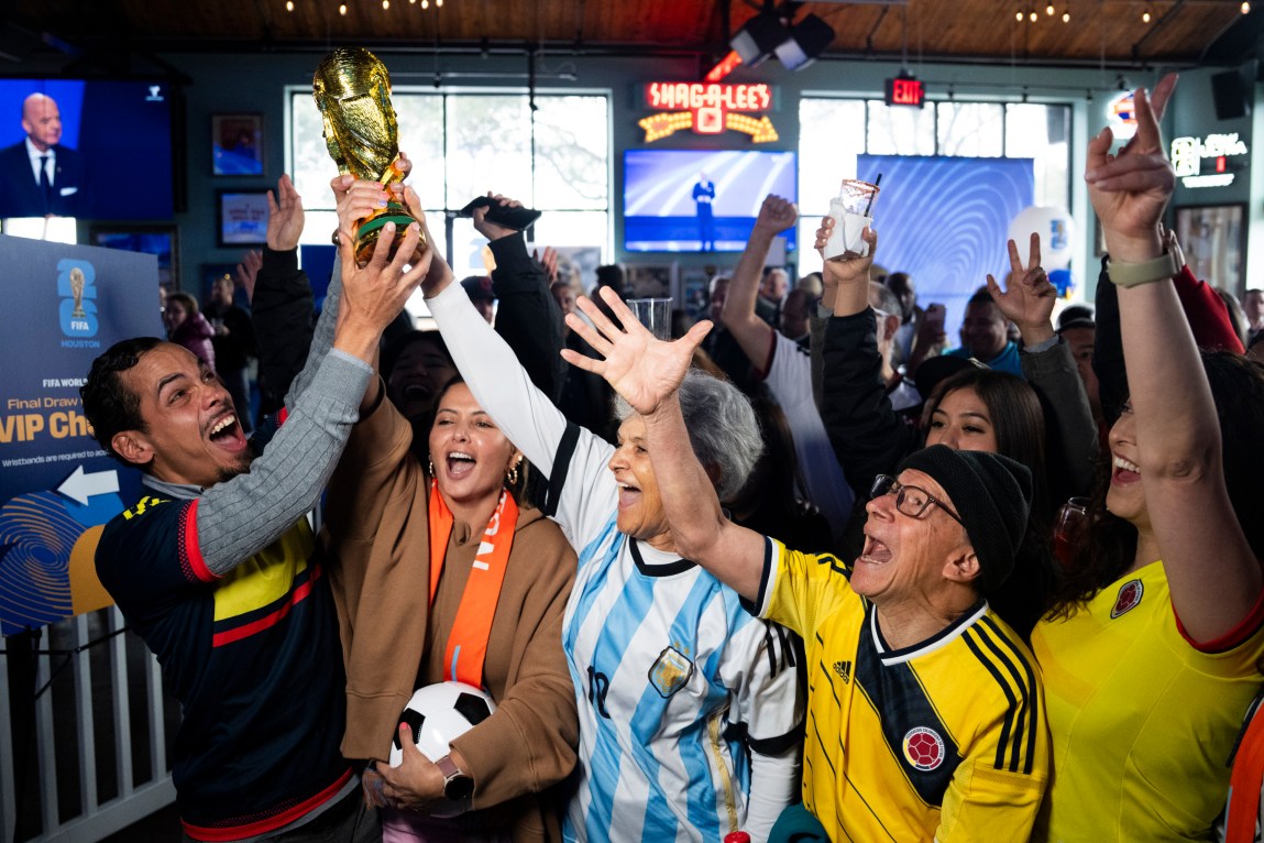 Fans hold up a soccer trophy while shouting and raising their arms in the air in a dark bar while illuminated by a flash.
