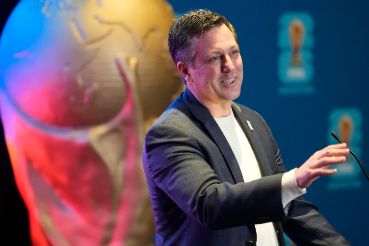 A man wearing a gray suit jacket and a white shirt speaks into a small microphone while standing in front of a large, gold soccer trophy.