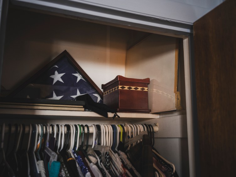 A small wooden box sits on a shelf inside a closet, next to an American flag and above clothing on hangers.