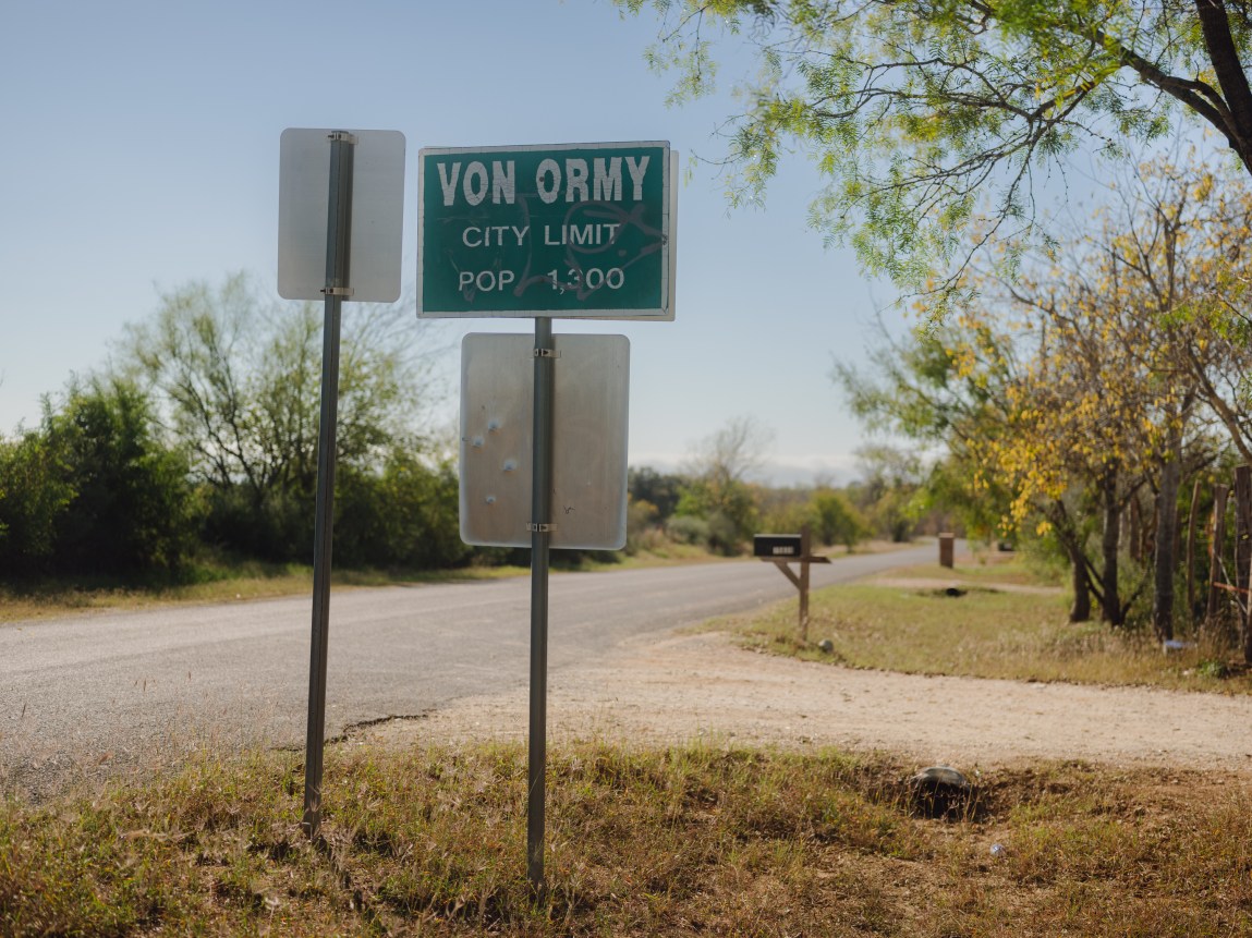 A small green sign on a post stating, “Von Ormy city limit” and “ pop 1,300,” alongside the silver backs of two other signs, on an empty country road lined with low trees and shrubs.