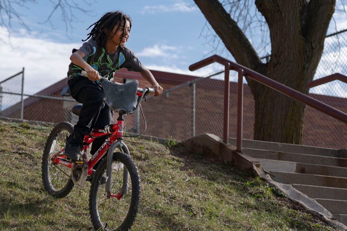 A child rides a red bike down a grassy hill, next to concrete stairs and a tree.