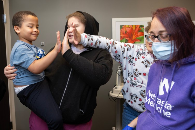 A teenager wearing a black hoodie holds and high-fives a boy wearing a blue shirt and black pants. Another child wearing a sweatshirt with peppermints, candy canes and mugs of hot chocolate also gives him a high-five. Off to the right, a woman wears gloves and a surgical mask, along with a sweatshirt that says Nebraska Medicine.