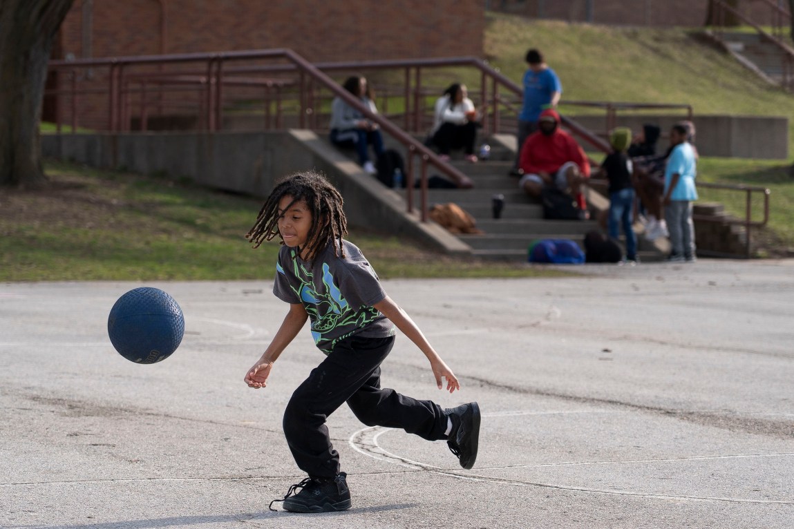 A child wearing a T-shirt, black pants and sneakers runs after a blue ball. Behind him are people sitting on a flight of steps leading out of a brick building.