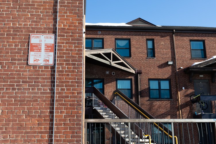 A brick building with stairs and windows reflecting a blue sky. A permit parking sign hangs on a wall with a notice saying vehicles without permits will be towed.