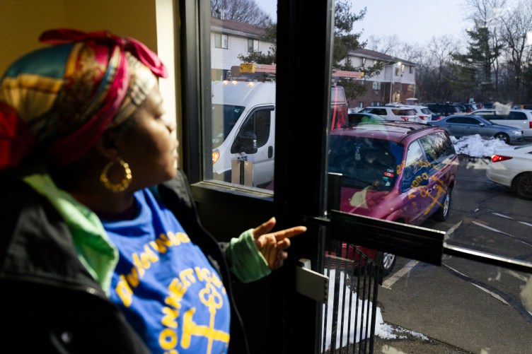A woman wearing a headscarf and gold hoop earrings stands inside pointing toward a glass door. A parking lot with cars and piles of snow is outside the door.