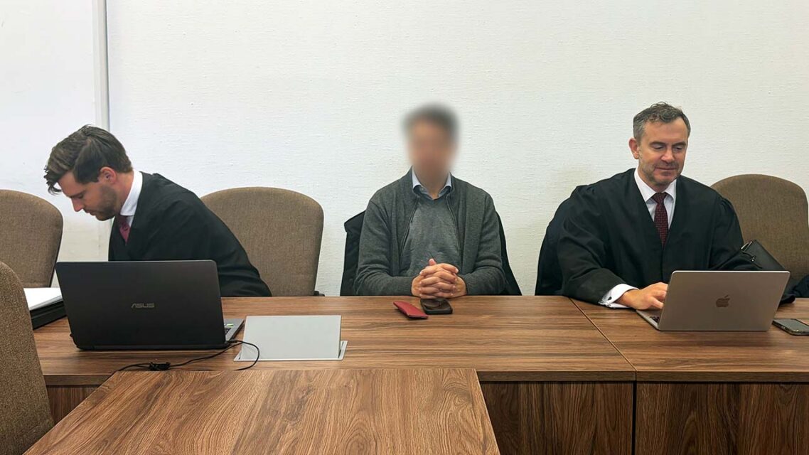 A man with a blurred face sits between two lawyers with laptops in a courtroom.