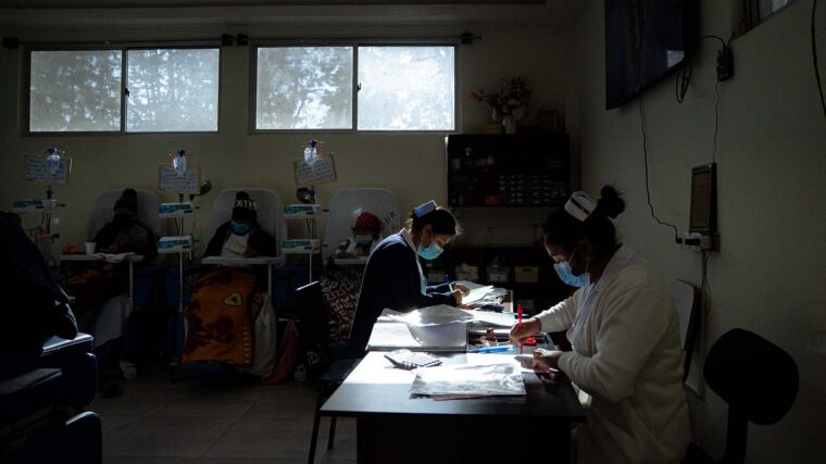 Photo of three patients sitting in chairs with IV drip bags, with nurses doing paperwork in the foreground.
