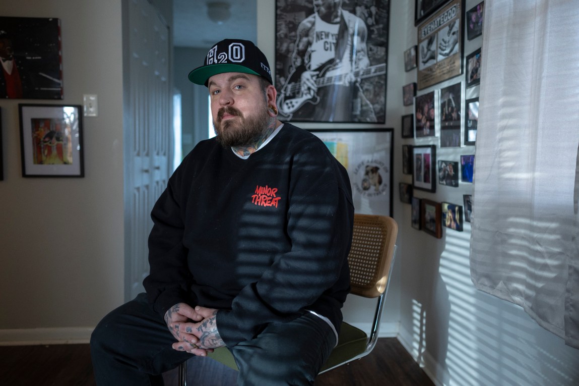 A man with a beard and tattoos on his hands wearing a baseball cap and sweatshirt, sitting on a chair in a room decorated with small framed photos.