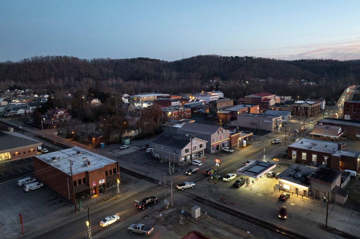 An aerial view of cars driving through a town with one-story buildings. Tree lined hills surround the town.