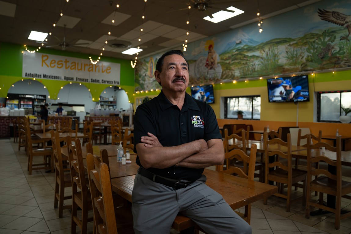 A man sits with his arms crossed on a table in an otherwise empty restaurant.