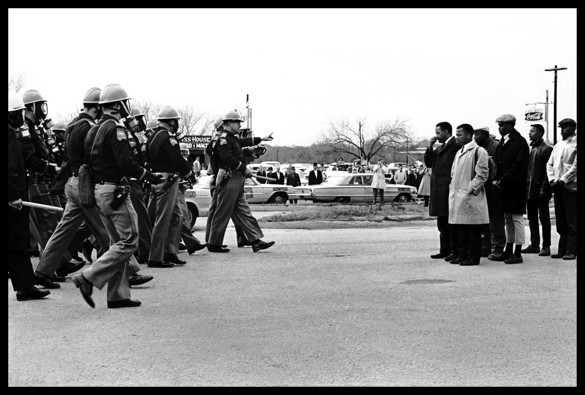 A black-and-white photograph shows a line of police officers advancing from the left side with billy clubs drawn and a group of Black men standing together on the right side. A crowd of people look on in the background.