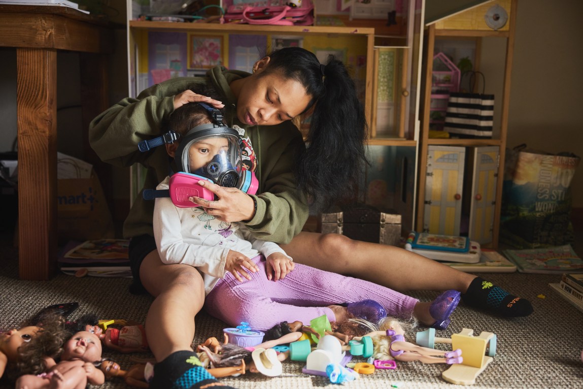 A woman and her young daughter sit on a living room floor, in front of a doll house and surrounded by dolls. The woman is putting a gas mask on the girl.