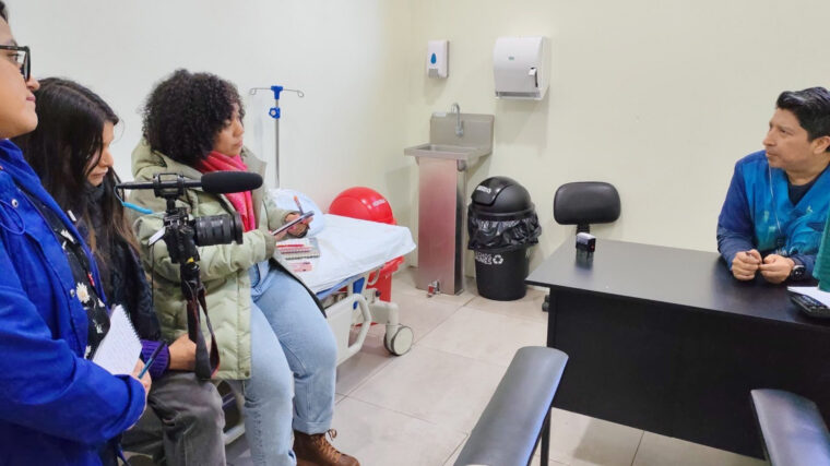 Three journalists with camera equipment interview a doctor behind a desk