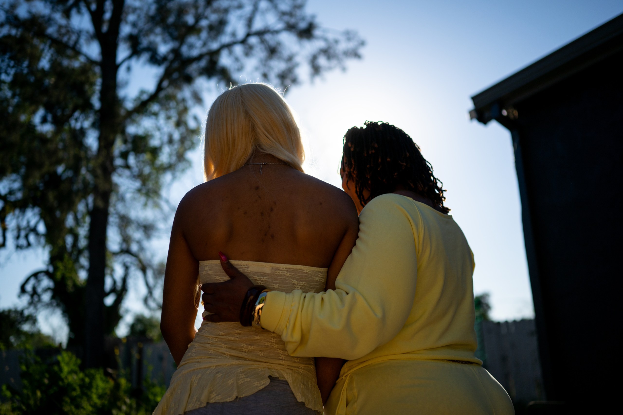 A 17-year-old girl and a woman stand outside with their backs to the camera. The woman rests her hand on the girl’s back in an embrace.