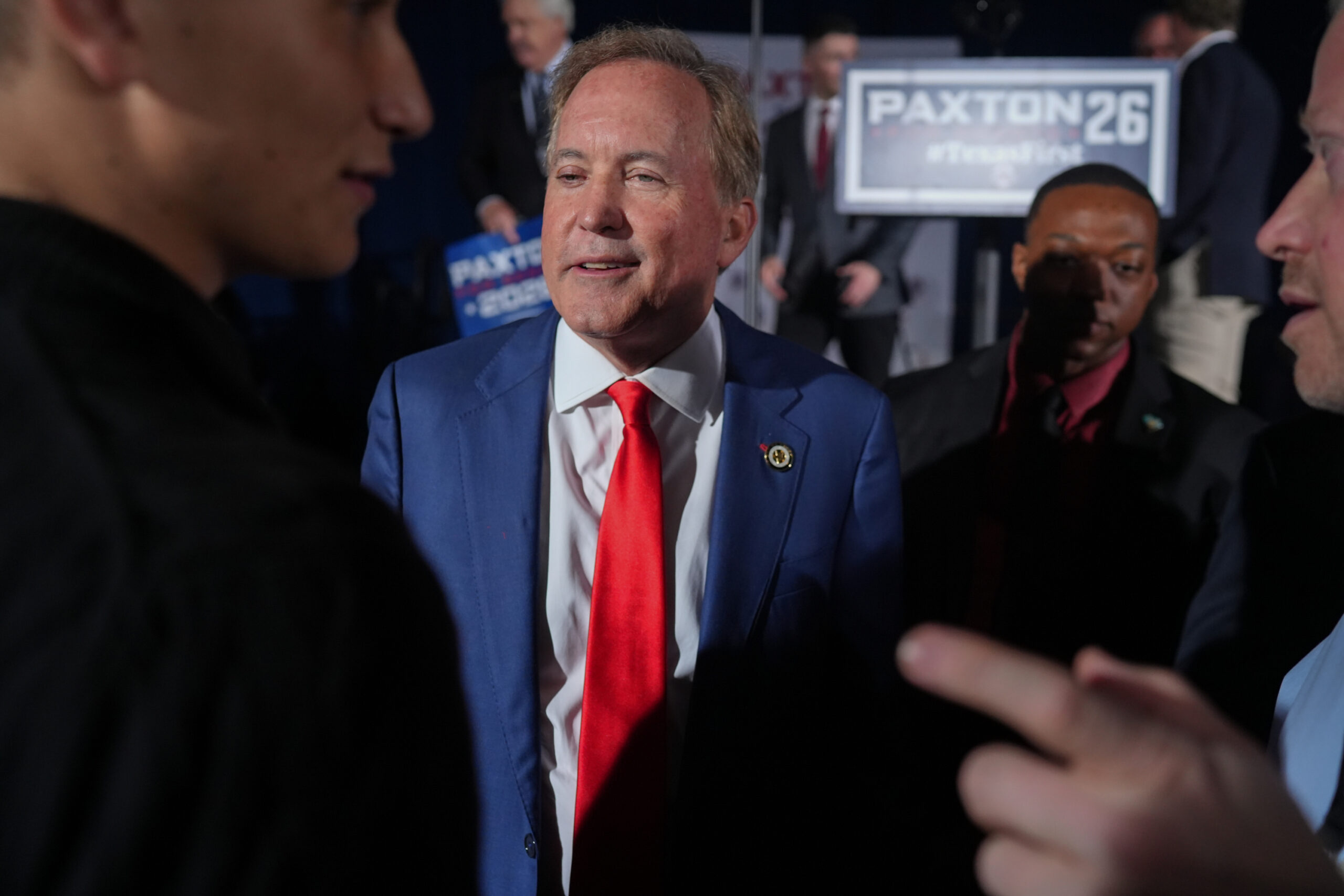 Ken Paxton, wearing a blue suit jacket and bright red tie, amid a crowd of people. In the background, a campaign sign reads “PAXTON 26 #TexasFirst.”