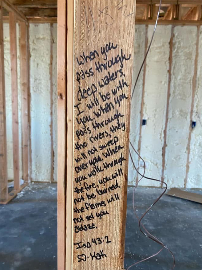 A wooden stud in a house has the words written on it: “When you pass through deep waters, I will be with you. When you pass through the rivers, they will not sweep over you. When you walk through the fire, you will not be burned, the flames will not set you ablaze.”