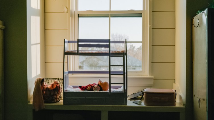 A doll lies in a doll bed, lit by a window behind it, with several household items around it.
