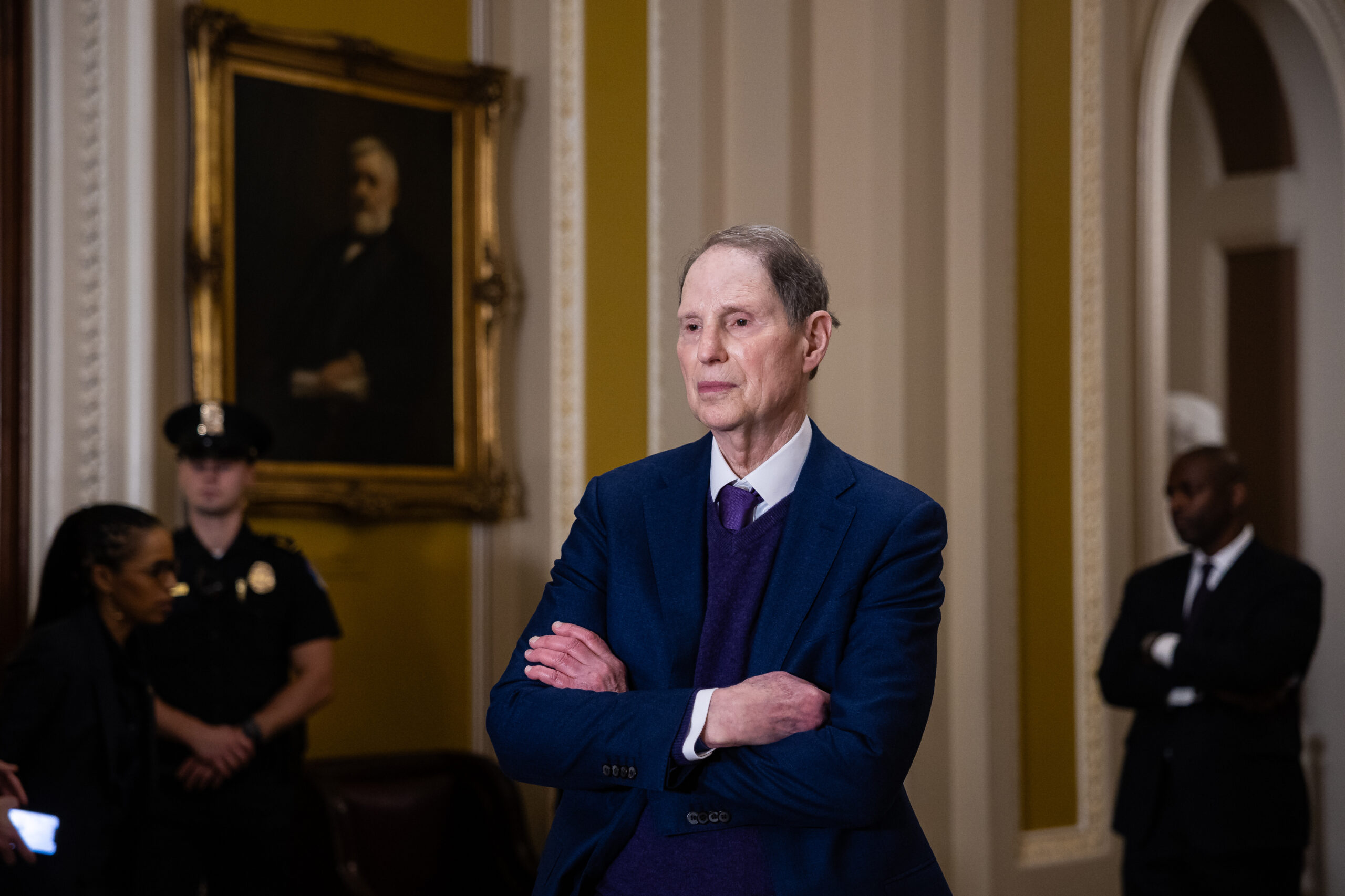 A man with gray hair wears a navy suit and tie and crosses his arms. In the background are three people, including a police officer and a man also crossing his arms wearing a black suit and white shirt. They are all standing in a room with an ornately framed portrait and gold-and-white walls with curved archways.