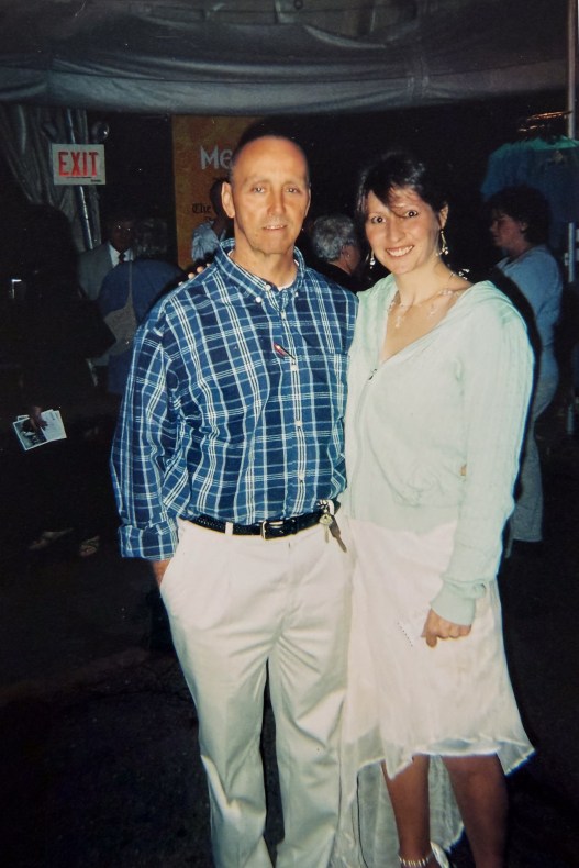 A man and a woman stand close together and look at the camera. There is a crowd of people in the background.