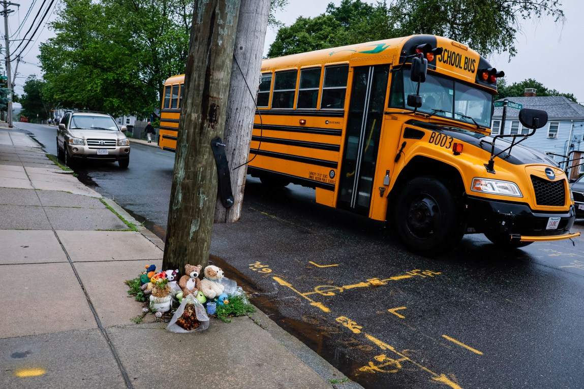 A yellow school bus on a city street next to a sidewalk memorial made up of stuffed animals and flowers.