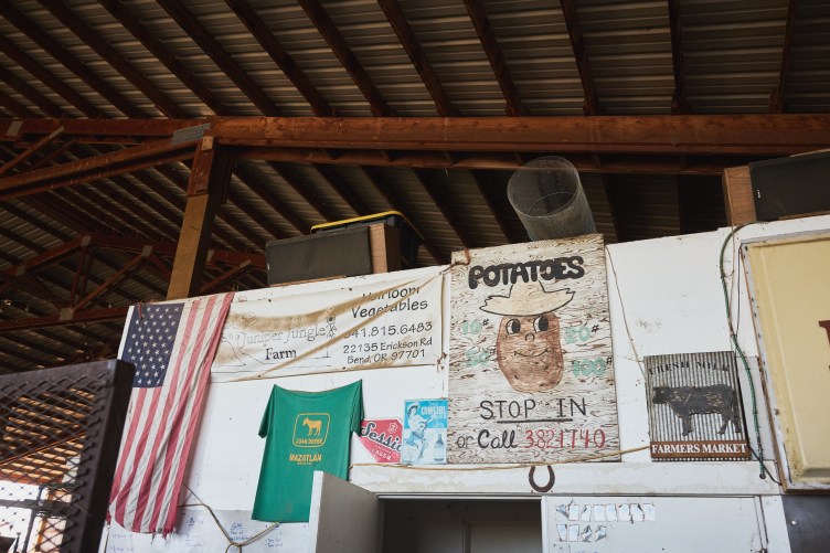 A wall in the interior of a barn with numerous objects hung on it, including a faded American flag, a T-shirt that says “Juan Deere” with a silhouette of a donkey, old advertising posters, and a board that reads, “Potatoes, Stop In” and has a drawing of a potato with a smiling face.