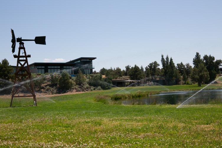 Sprinklers shoot into the air to water a large lawn surrounding a pond and a small windmill. In the background is a house on a hill that overlooks the property.