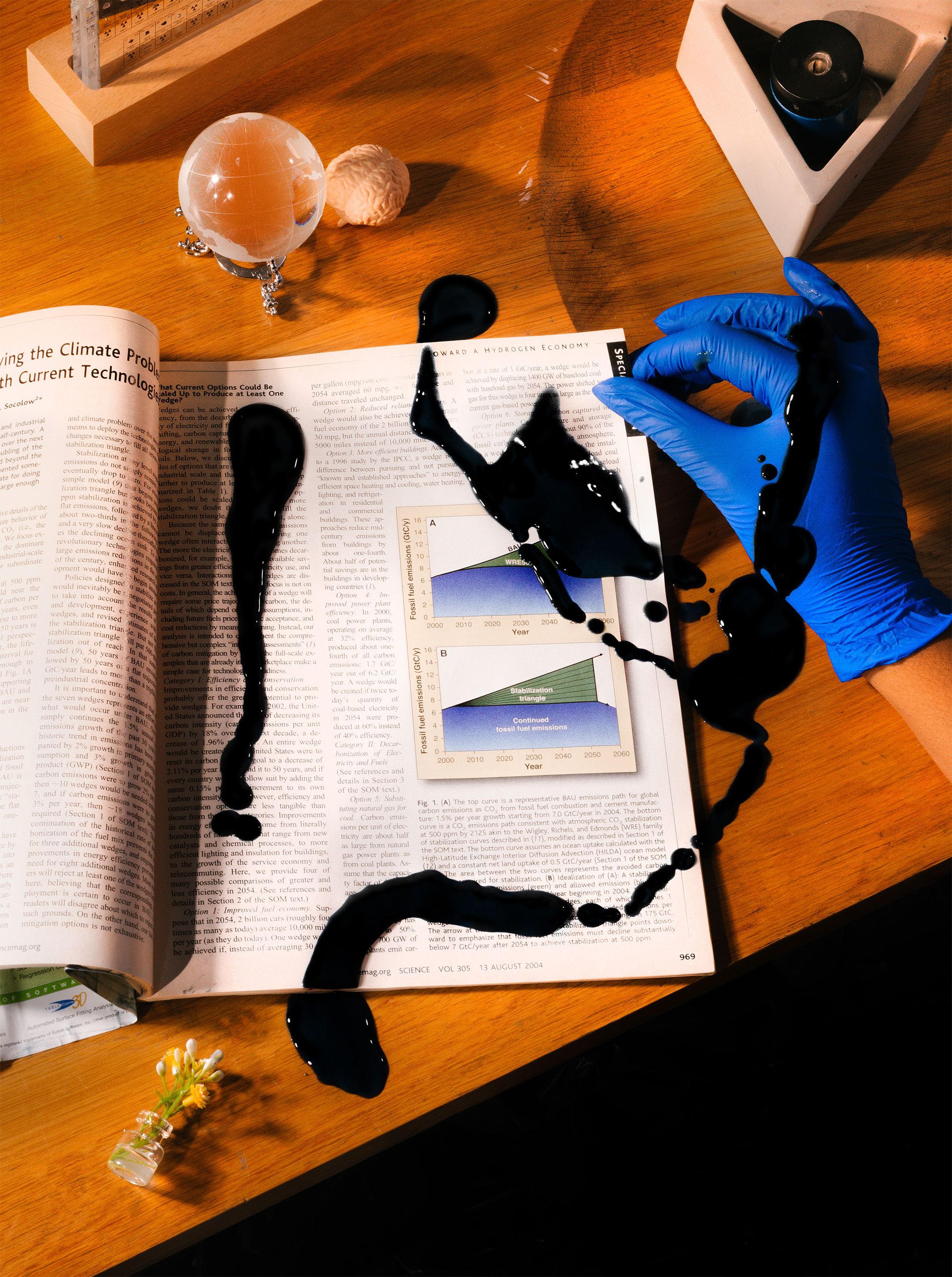 A science magazine is open on a wooden desk surrounded by trinkets in the shape of a globe, a brain and flowers. A hand with a blue medical glove is lifting up a page with oil splattered over it.